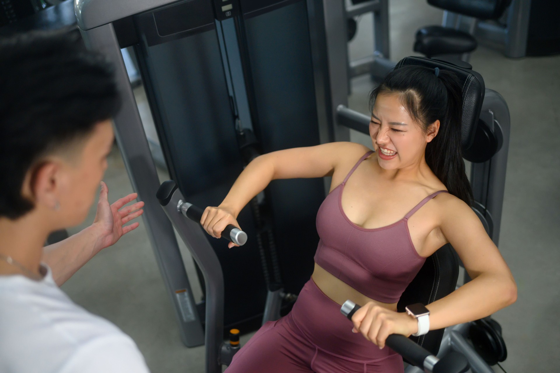 Determined woman pushing through chest press exercise on machine with a trainer nearby for guidance